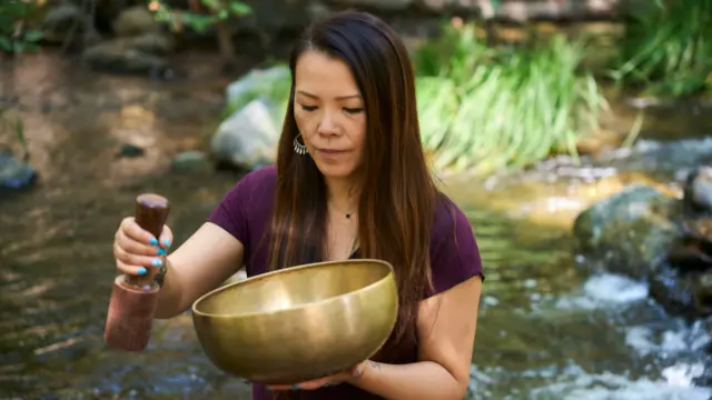 Woman using a Tibetan singing bowl for healing and inner peace beside a flowing stream