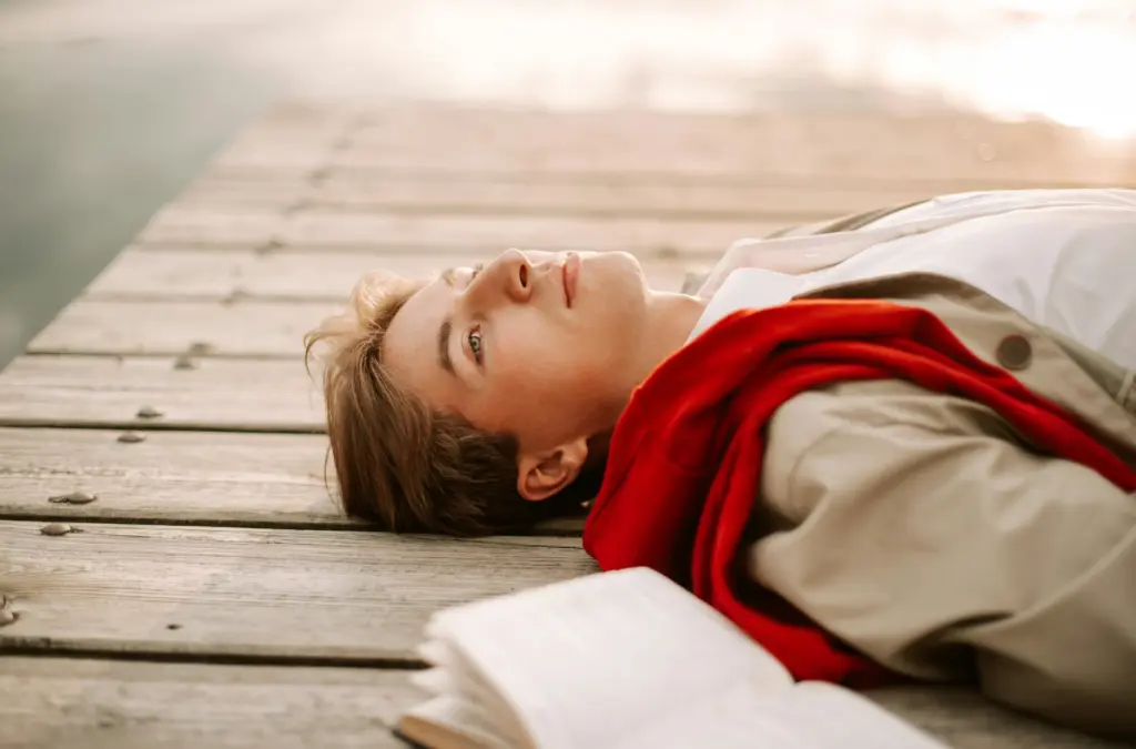 Person lying on a wooden deck near water, reflecting with an open book beside them