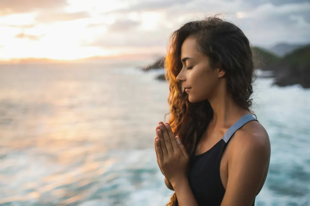 Woman standing by the ocean at sunset with eyes closed and hands together, symbolizing peace, faith, and inner calm.