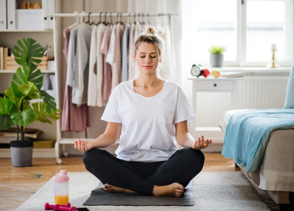 Woman practicing mindful breathing at home, sitting cross-legged on a yoga mat with eyes closed and calm expression.