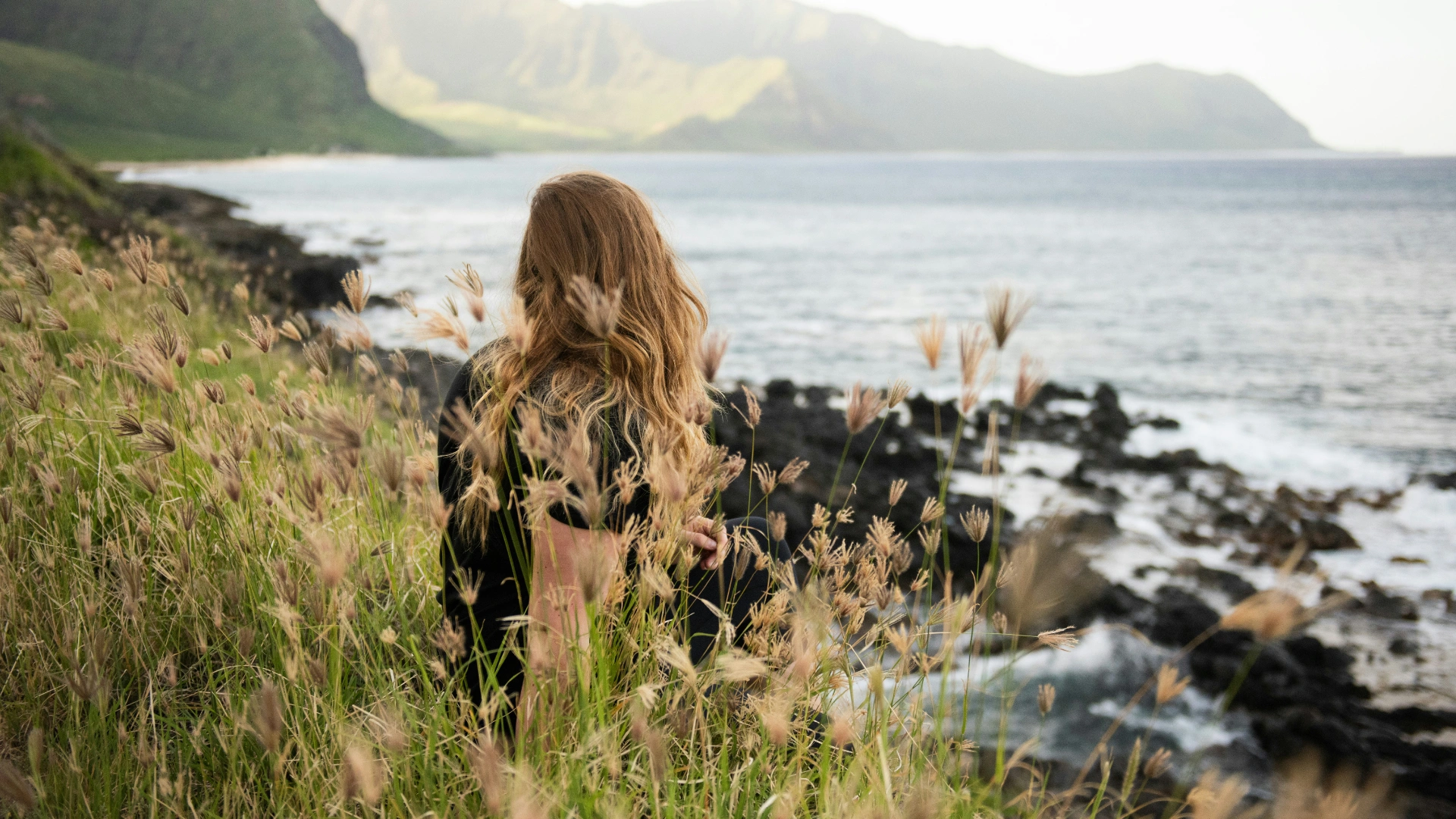 Woman sitting in tall grass overlooking the ocean, symbolizing calm reflection, healing, and emotional resilience.
