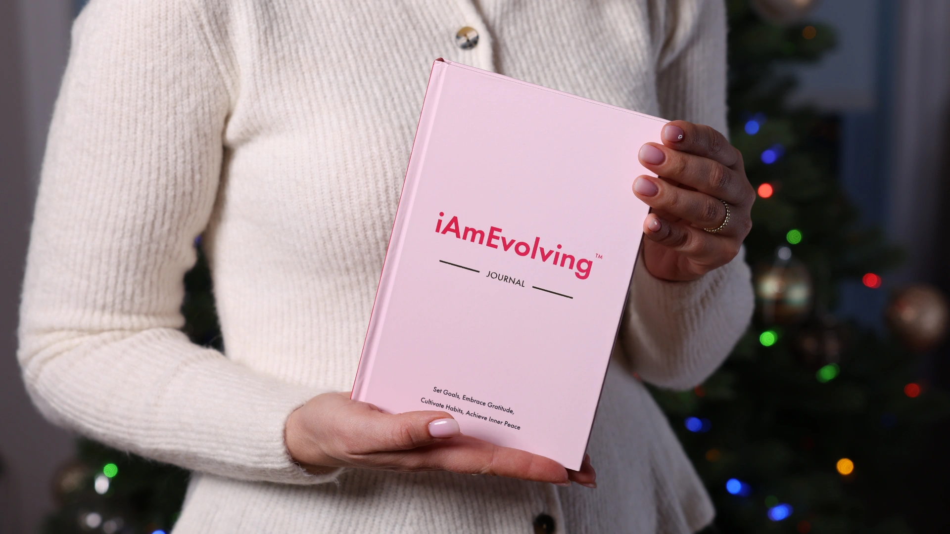 Person holding the iAmEvolving Journal near a softly lit holiday tree — reflecting on gratitude through the seasons.