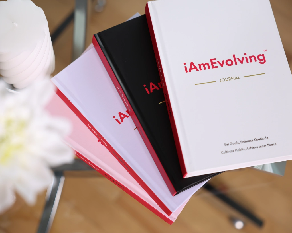iAmEvolving Journal stacked in all four hardcover colors: white, black, misty rose, and lavender on a glass table with flowers