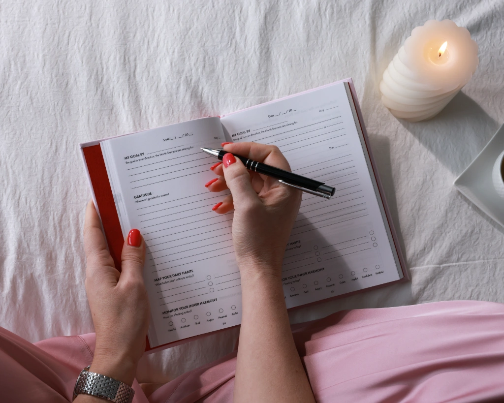 Woman writing in the iAmEvolving Journal daily page showing goal, gratitude, habits, and inner harmony sections, with candle and coffee on bed