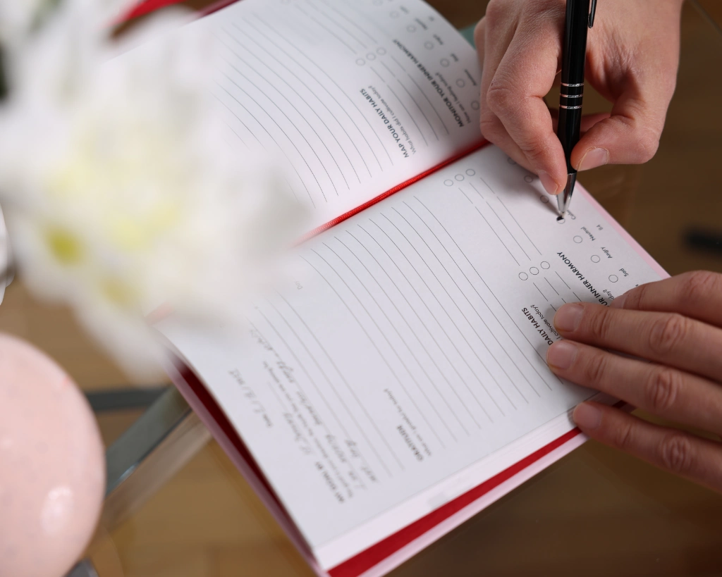 Hands writing in the iAmEvolving Journal showing daily habits and inner harmony prompts, with flowers on a glass table